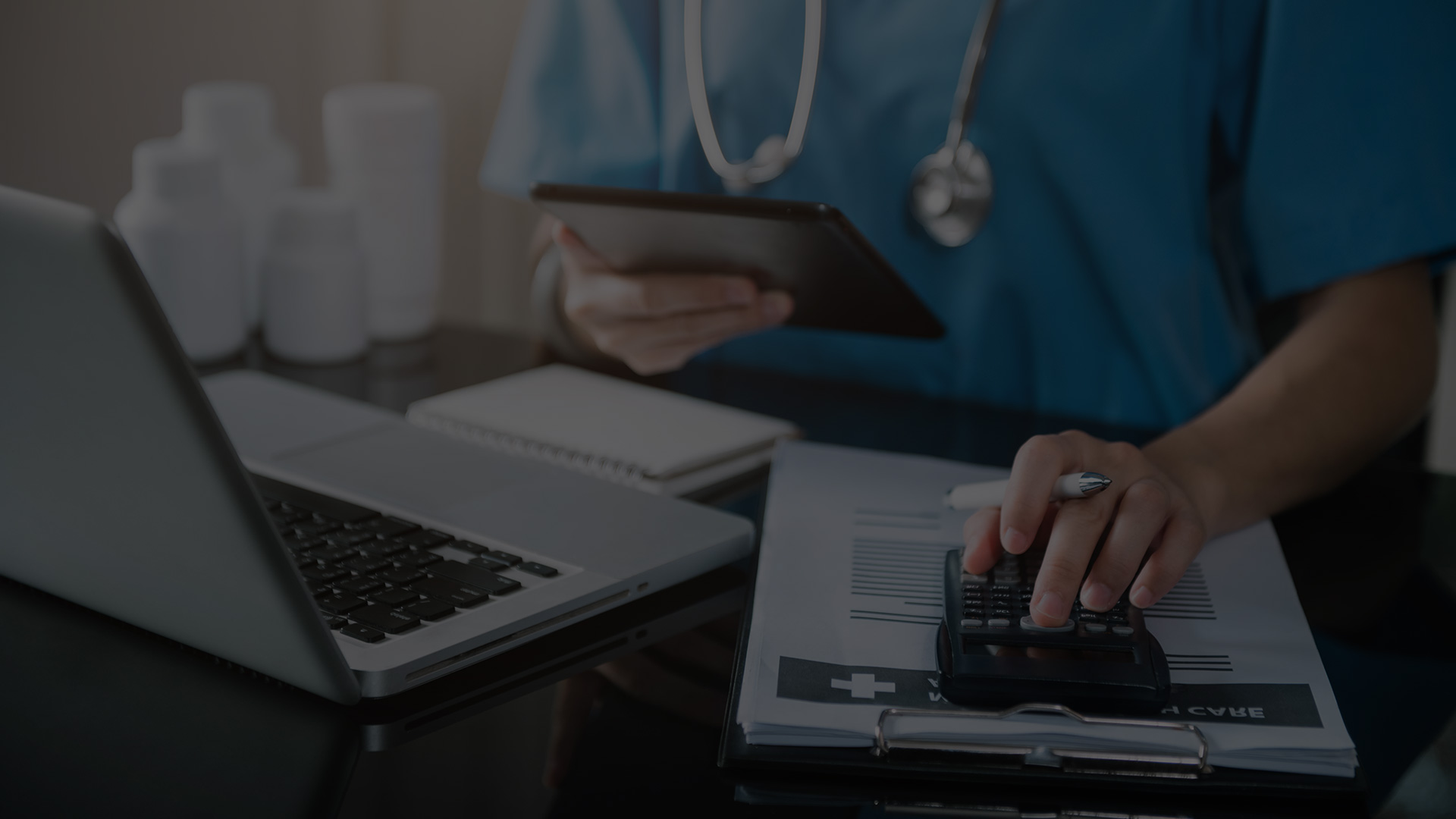 Healthcare professional looking at papers and laptop while holding a tablet and using a calculator