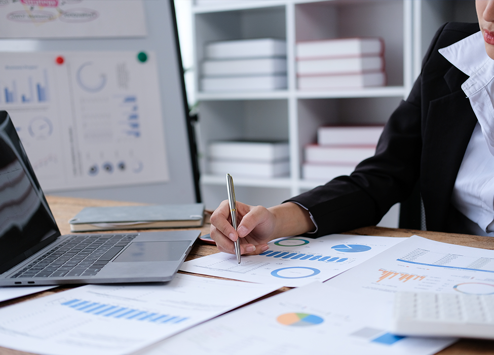 Papers are scattered around a laptop while a person writes notes on paper with graphs 