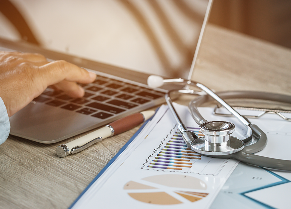 Denial Management in Healthcare | Close up of person working on laptop with papers and a stethoscope resting next to them